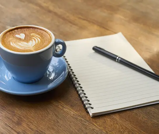 Hände mit Kaffeetassen, Notizbuch und Smartphone an einem Holztisch im Freien
